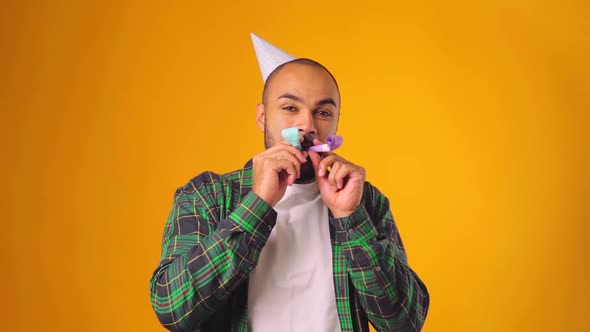 Happy African American Male in Party Cone Blowing Party Blower Against Yellow Background