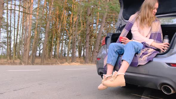 Young Woman Sits in Trunk of Car Against Backdrop of Pine Grove on Sunny Autumn or Spring Day