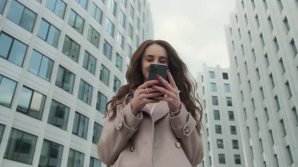 Cheerful young Caucasian woman standing in city street and texting on mobile phone with smile.