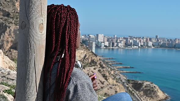 African Woman Sitting on the Sea and City Background and Listening to the Music