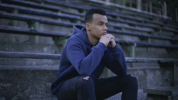 Upset Afro-American Teenager Sitting on Tribune, Devastation and Poverty Around