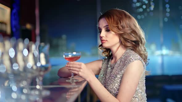 Beautiful Young Woman Drinking Cocktail Sitting on Bar Looking at Camera at Disco Party