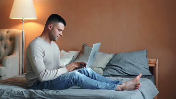 Casual Young Barefoot Freelancer Male Chatting Working Using Laptop Pc Sitting on Bed