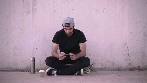 Man sitting on skateboard using phone and drinking coffee