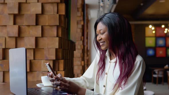 Young African Woman Using Phone While Sitting in a Cafe with a Laptop