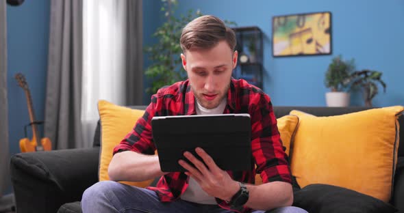 Serious Millennial Male Sit on Couch Focus Attention on Digital Pad Screen Read Electronic Book