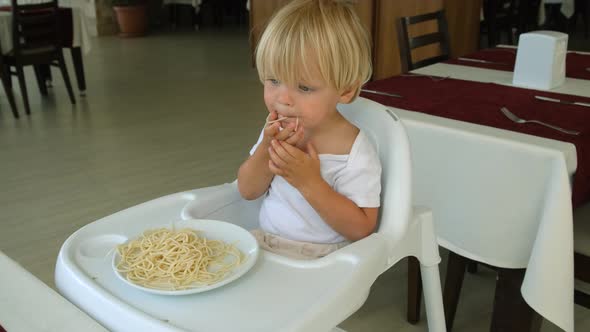 Baby Boy Eats Spaghetti Sitting in a Baby Chair in Restaurant