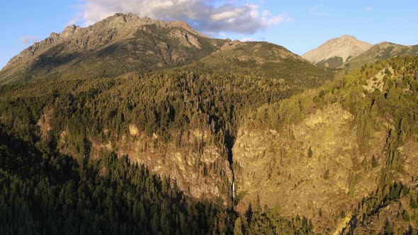 Aerial dolly out over Corbata Blanca thaw waterfall hiding between pine tree forest mountains at sun