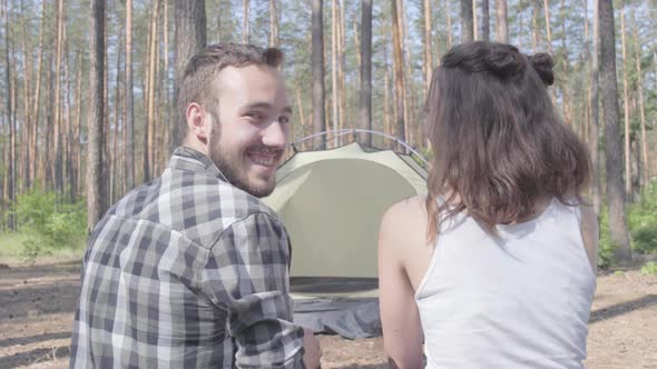 The Young Man and Woman Sitting Near Each Other with Backs To Camera in Forest. The Tent on the