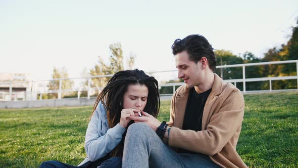 Happy Young Hipster Couple Sitting on Grass and Smoking Marijuana Joint in Park