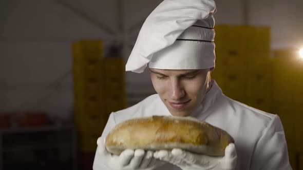 Happy Baker in Uniform Sniffs Loaf of Bread Has a Pleasure and Smile at Camera