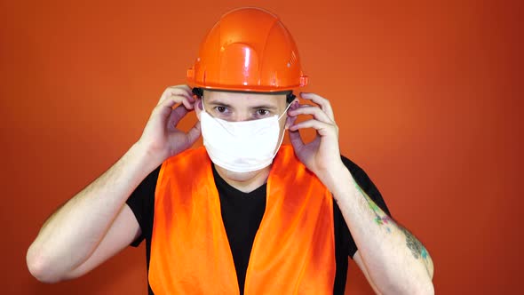 Male Construction Worker in Overalls Putting on Medical Mask on Face on Orange Background