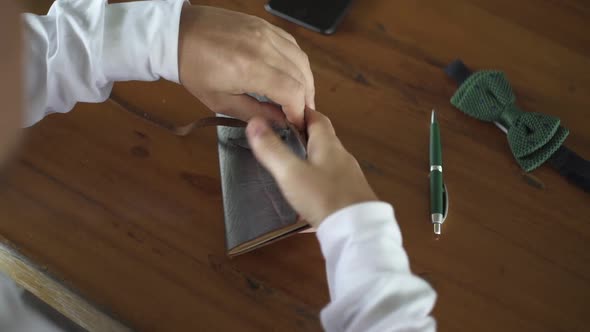 Groom finish writing vow, close leather notebook. Accessories, phone, bow-tie, pen on wooden table.