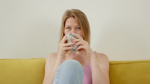Beautiful Cute Woman in Pink Top Sitting on Yellow Sofa at Home Drinking Hot Fragrant Coffee or Tea