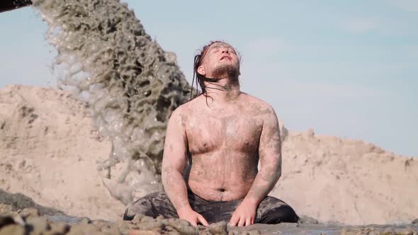 Funny Fat Man with Long Hair Sits in the Mud. Against the background pouring Black Dirty Water Pipe