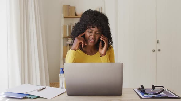 African american woman using laptop and talking on smartphone while sitting on her desk at home