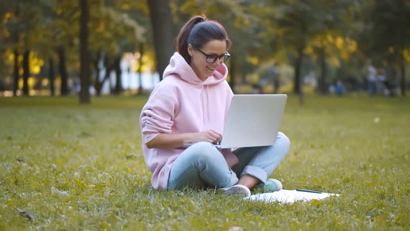 Cheerful Young Woman in Specs Using Pc Sitting in Park Preparing for Exams