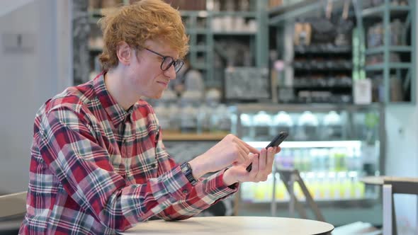 Unhappy Young Redhead Man with Loss on Smartphone in Cafe