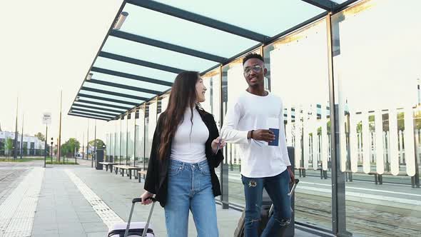 Two Mixed Race Friends with Suitcases on Wheels and Documents in their Hands Waiting for Shuttle Bus