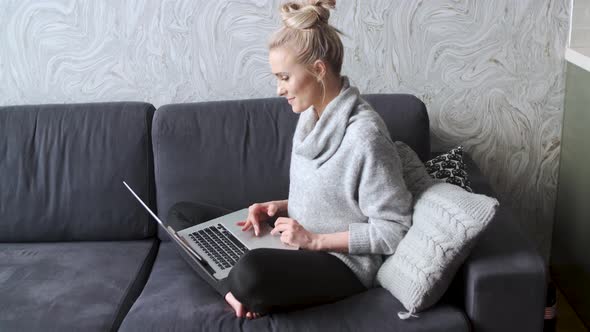 Cheerful Young Blond Woman Sitting on Couch in Living Room and Using Laptop