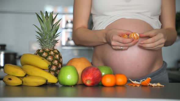 Pregnant Woman Eating a Tangerine in Home Kitchen with Different Tropical Fruits