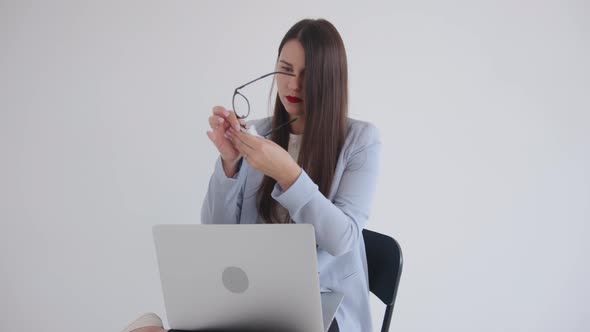 A Beautiful Young Business Woman Sits on a Chair with a Laptop on Her Lap and Wipes Her Glasses with