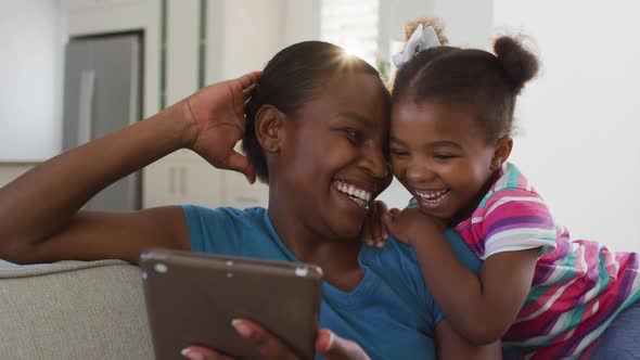 Happy african american mother and daughter sitting on sofa using digital tablet and laughing