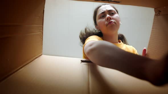 A young woman opens the cardboard box in frustration, taking out books and perplexed indignation.