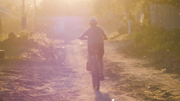 Boy Rides a Bicycle in the Sunset Light Along the Village Road