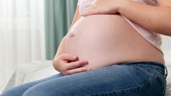 Closeup of Beautiful Pregnant Woman with Big Belly Sitting on Bed and Touching Her Abdomen