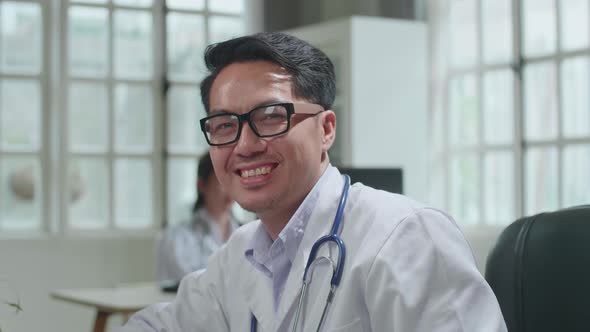 Asian Man Doctor Is Using Desktop Computer, He Turns And Smiles To Camera In Workplace