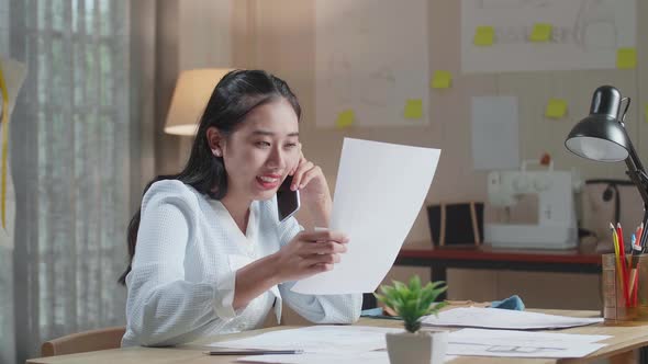 Asian Woman Designer Talking On Smartphone While Looking At The Layout Bond In Hand At The Office