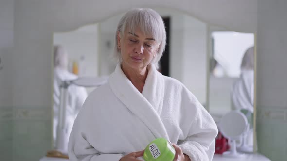 Sad Caucasian Middleaged Greyhaired Woman Standing in Bathroom with Aloe Cream Looking at Camera