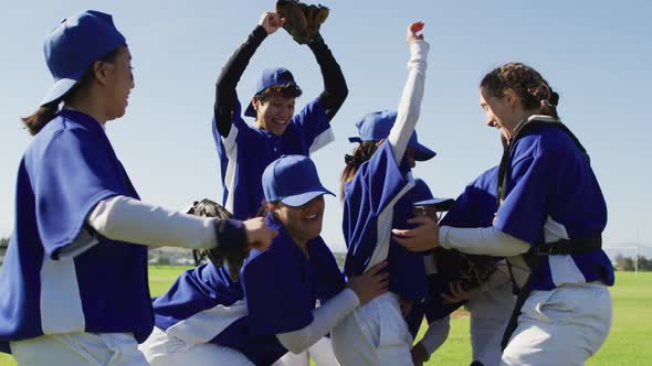 Happy diverse team of female baseball players celebrating after game, lifting up one player