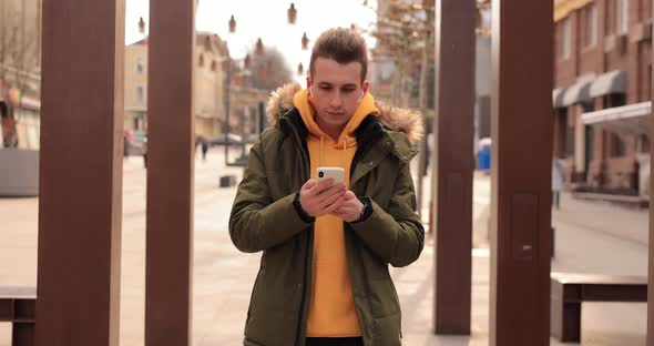 Man Walks in a Winter City with Wireless Earbuds in His Ears and Smartphone