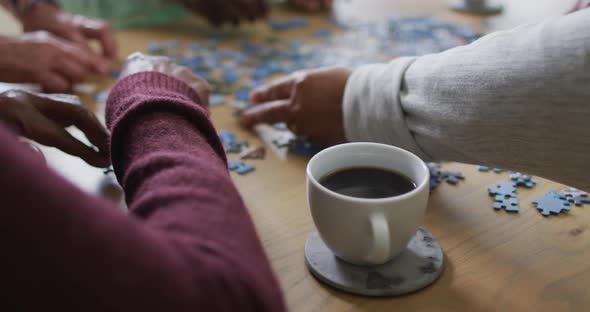 Hands of group of diverse senior friends drinking coffee and doing puzzle at home