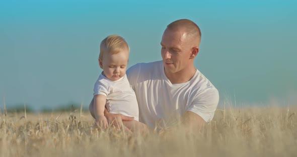 Young Man is Sitting in a Wheat Field and Holding a Baby in His Arms They are Smiling