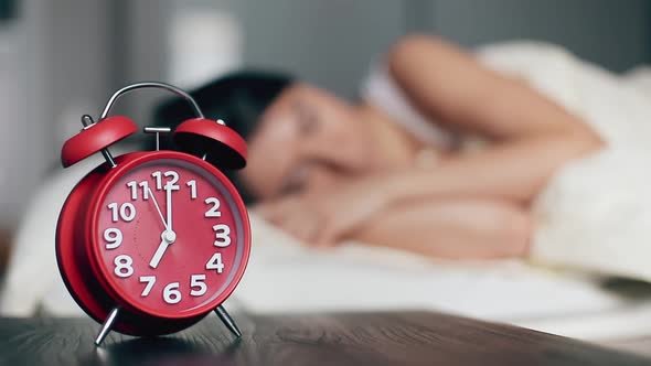 The Alarm Clock on a Bedside Table Against the Background of the Sleeping Woman