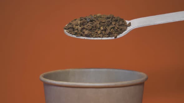 Coffee Falls in Slow Motion From a Wooden Spoon Into a Craft Paper Glass on an Orange Background