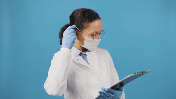 Woman Doctor with Clipboard Putting on Protective Medical Mask Before Appointment with Patient