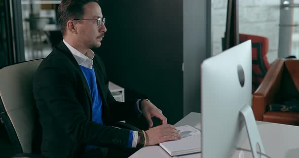 Businessman in Glasses Sits at Computer in Office and Writing It Into a Notebook