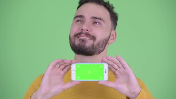 Face of Happy Young Handsome Bearded Man Thinking While Showing Phone