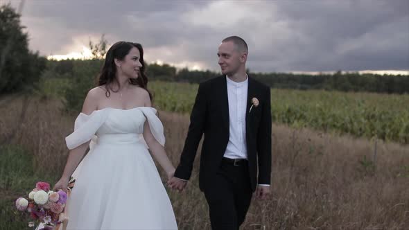 The Bride with a Bouquet of Dahlias Holds the Groom's Hand and They Walk Across the Field Looking