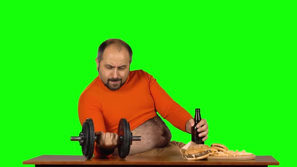 Overweight Caucasian Man Sits at a Table with Junk Food in One Hand Holds Dumbbell in Second Bottle