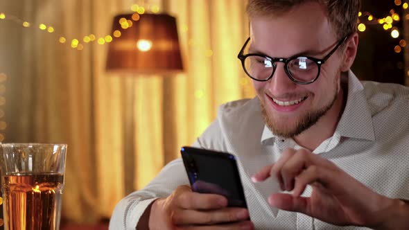 Handsome Bearded Man Using a Smartphone and Smiling While Drinking Beer in Pub