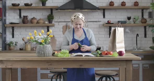 Female Caucasian Retiree Peeling Cucumber with Kitchen Knife and Looking Into Recipe Book. Senior