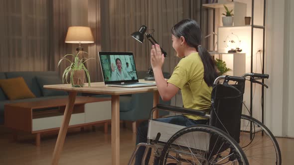 Asian Kid Girl Sitting In A Wheelchair While Video Call With Doctor On Laptop Computer At Home