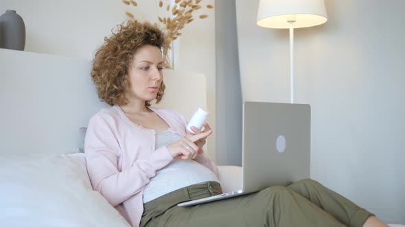 Pregnant Woman Using Laptop And Reading Label On Medicine Bottle At Home