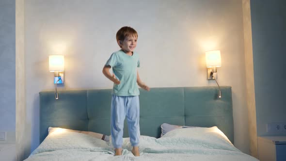 Happy Cheerful Toddler Boy in Pajamas Jumping Up High on Big Bed at Night Before Going To Sleep