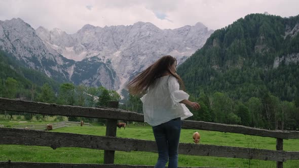 Young Woman is Jumping and Whirling on Meadow Near Alps and Farm